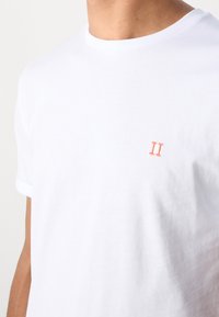 Weißes T-Shirt mit Kurzarm und Rundhalsausschnitt, mit einer kleinen, orangen römischen Zahl II auf der linken Brust bestickt, getragen von einer Person.