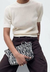 Cremefarbenes Stricktop mit kurzen Ärmeln, kombiniert mit dunkelbrauner Hose. Hält eine schwarz-weiß gestreifte Clutch mit Henkel in der Hand.