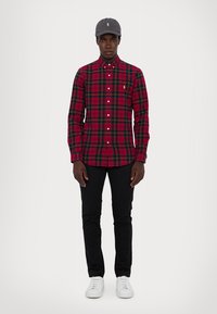 Κόκκινο και μαύρο καρό πουκάμισο με γιακά που κουμπώνει, σε συνδυασμό με μαύρο παντελόνι και λευκά αθλητικά παπούτσια. Γκρι καπέλο με λογότυπο ολοκληρώνει την εμφάνιση.