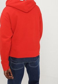 Persona vistiendo una sudadera con capucha roja vibrante y jeans azul oscuro, mostrada de espaldas contra un fondo claro y liso.