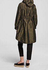 Cappotto impermeabile leggero di colore verde oliva, con cappuccio, vita elastica e una stampa logo sottile. Indossato sopra pantaloni neri aderenti e scarpe espadrillas.