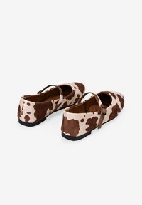 Scarpe in stile Mary Jane per bambini con motivo a macchie di mucca marrone e bianco e cinturini neri con fibbia, su uno sfondo bianco.