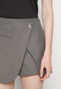 Jupe portefeuille grise en tissu texturé, avec un design épuré, une fermeture latérale à boutons et une coupe ajustée, présentée avec un haut noir.