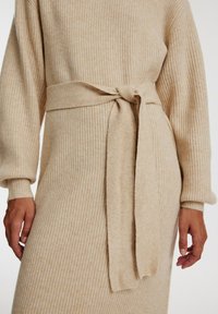 Beige Strickkleid mit gerippter Textur, das einen gebundenen Gürtel in der Taille und lange, leicht puffige Ärmel hat.