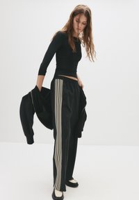 Schwarzes Sportoutfit mit einem taillierten Langarm-Oberteil und lockeren Jogginghosen, die entlang der Seiten hellfarbige Streifen haben.