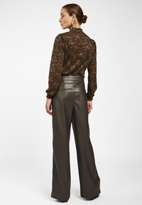 Frau mit braunem Haar im Dutt trägt eine durchsichtige, dunkelbraune Bluse mit Blumenmuster und hochgeschnittene, weit geschnittene, dunkle Lederhosen, steht vor weißem Hintergrund.