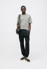 Γκρι βαμβακερό t-shirt, μαύρο παντελόνι jogging και μαύρα αθλητικά παπούτσια με λευκά κορδόνια. Το μοντέλο κρατά μια μαύρη τσάντα, ενώ στέκεται σε λευκό φόντο.