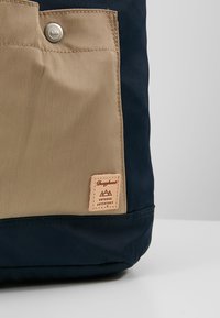 Plecak z granatowym korpusem, beżową kieszenią z przodu i małą skórzaną metką z logo "Doughnut". Wykonany z trwałego materiału nylonowego.