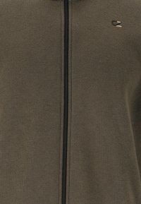 Oliivinvihreä vetoketjullinen huppari pehmeästä kankaasta. Varustettu mustalla vetoketjulla ja pienellä logolla rinnassa. Yksinkertainen muotoilu, jossa on sileä pinta.