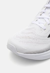 Chaussure de sport blanche avec un dessus en maille, présentant des motifs de ventilation intricats, des lacets blancs plats et une semelle en caoutchouc avec un minimum de marquage.