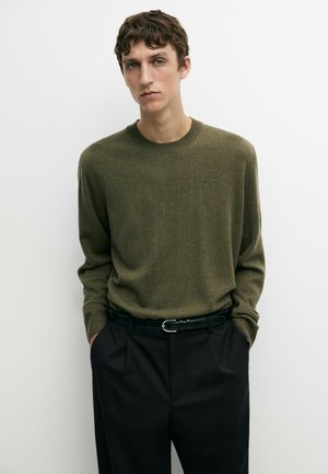 Junger Mann mit kurzen braunen Haaren trägt einen olivgrünen Pullover, der in hoch taillierte schwarze Hosen mit einem schwarzen Gürtel gesteckt ist, steht vor einer weißen Wand.