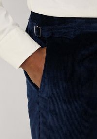 Tamno plave korduroy hlače s teksturiranom površinom, s bočnom džepom i malom metalnom kopčom na struku. Uparene s bijelom košuljom.