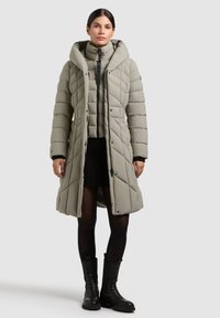 Light gray quilted puffer coat with oversized hood, zip closure, and front pockets, layered over a black knit dress and tights, paired with black boots.