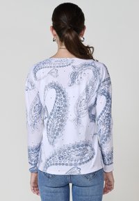 Mujer con cabello castaño en una coleta, usando pendientes de aro plateados, una camisa blanca de manga larga con patrones de paisley azules y jeans azules, de espaldas.