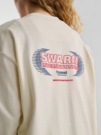 Persona bionda che indossa una felpa color crema con i loghi "SWARM INTERNATIONALS" e "hummel sportswear" stampati sul retro.