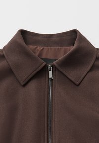 Braune Stoffjacke mit einem Kragen und einem silbernen Reißverschluss. Die Textur wirkt glatt mit einem leicht strukturierten Design.