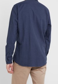 Man i en marinblå långärmad skjorta och beiga byxor, sedd bakifrån mot en enfärgad vit bakgrund.