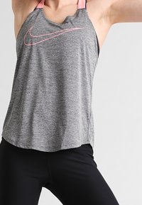 Débardeur de sport gris avec bretelles roses, arborant un grand logo Nike rose, coupe ample, fabriqué en matériau respirant.