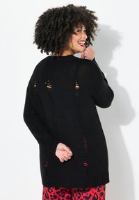 Schwarzer Strickpullover mit lockerem Schnitt, der ausgeschnittene Details auf dem Rücken und gerippte Bündchen hat. Kombiniert mit roten Leopardenmusterhosen.