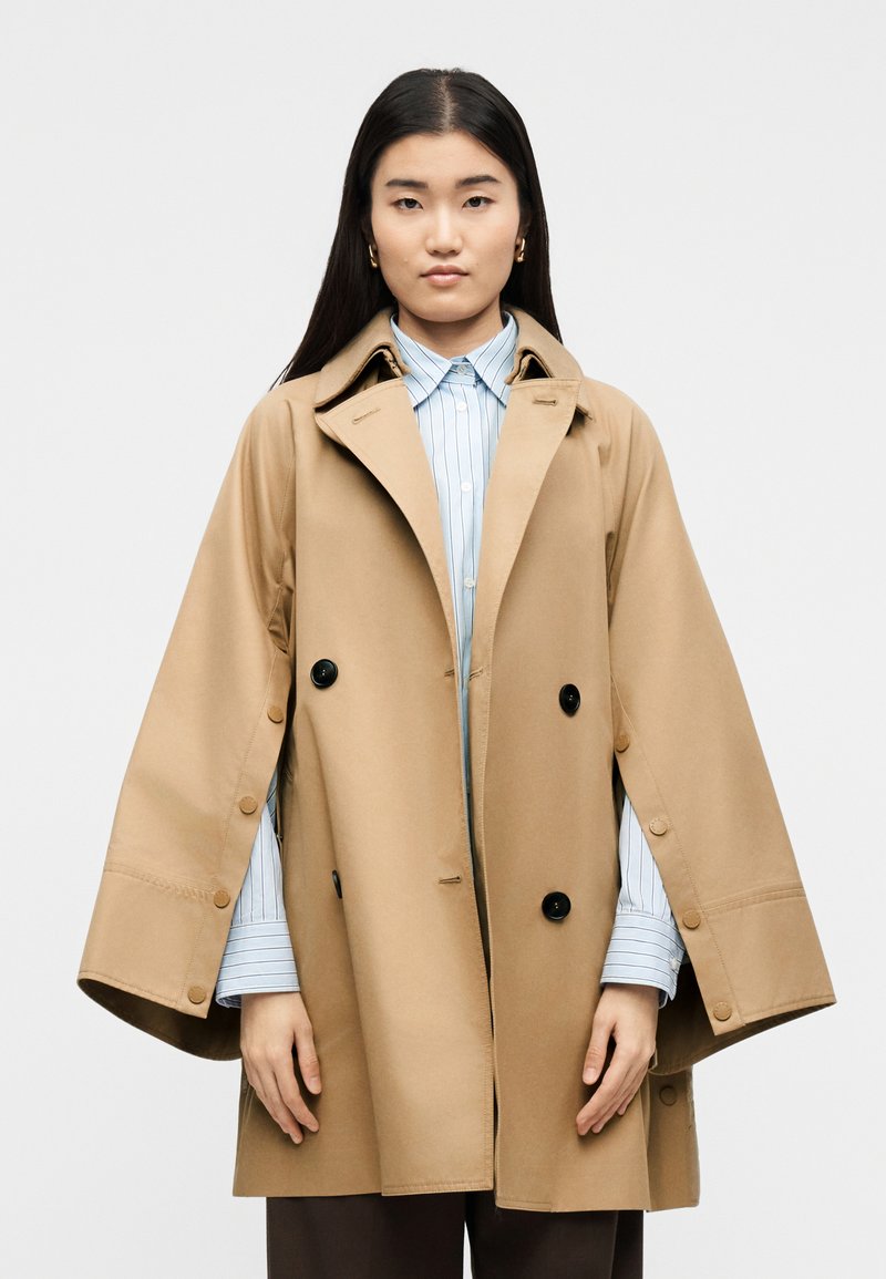 Γυναίκα που φοράει ένα διπλής σειράς μπεζ πανωφόρι-κάπα πάνω από μια μπλε ριγέ πουκάμισο με κουμπιά, όρθια μπροστά σε απλό λευκό φόντο.