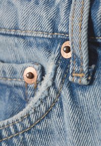 Svetlo modro jeansa tkanina z teksturiranim tkanjem, na kateri so bakrene zaponke in vidni šivi ob robovih ter odprtini žepa.