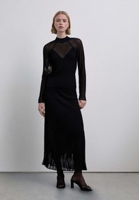 Mujer con top negro transparente de manga larga, falda midi negra con flecos en el dobladillo, sandalias negras de tacón y sosteniendo un bolso negro pequeño.