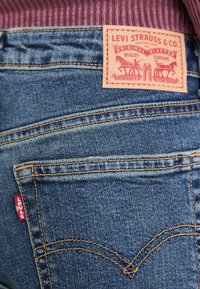 Vaqueros Levi's de mezclilla azul con una etiqueta trasera de cuero marrón que presenta un logo estilizado y costuras. Pestaña roja visible en el bolsillo.