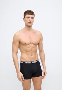 Homme torse nu aux cheveux bruns courts portant un boxer noir Puma, debout devant un fond gris clair uni.