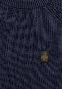 Tessuto lavorato a maglia blu navy con colletto a costine e una piccola toppa nera con un logo di cane giallo e testo nella zona del petto.