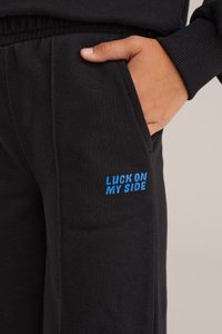 Mustad mustad püksid pehme tekstuuriga, küljetasku ja sinine tikand "LUCK ON MY SIDE" tasku lähedal.