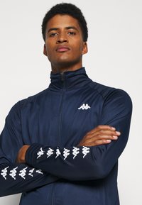 Tengerészeti zipzáras dzseki sima, könnyű anyagból. Magas gallérral és fehér logóval a mellkason, fehér mintákkal az ujjak mentén.