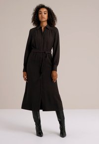 Robe noire à manches longues avec un devant à boutons, une ceinture à la taille et des poignets ajustés ; le tissu semble lisse avec une silhouette fluide.