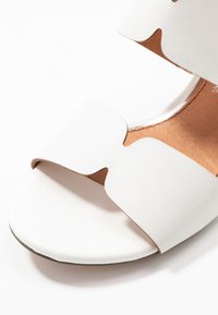 Sandal bianco con un ampio design della fascia ondulata, texture liscia, punta rotonda e soletta marrone chiaro. Suola piatta con dettagli sottili.
