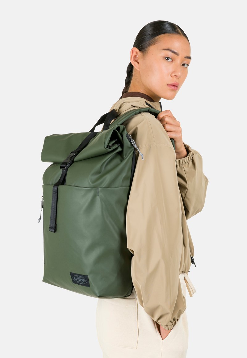 Jeune personne avec une queue de cheval tressée portant une veste beige et un grand sac à dos vert à rabat enroulable sur une épaule.
