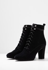 Botas de tobillo de ante negro con un alto tacón cuadrado, que presentan un diseño de cordones y ojetes metálicos. Textura suave, sin patrones visibles.