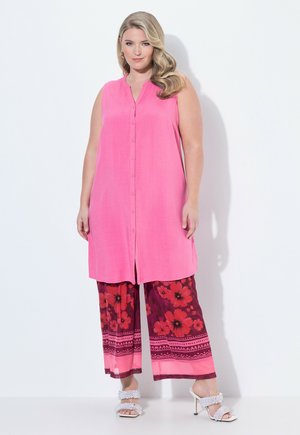 Kobieta o długich blond włosach, ubrana w różową bezrękawnikową tunikę na guziki, szerokie spodnie w kwiatowy wzór oraz białe sandały na wysokim obcasie.