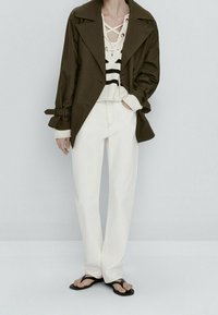 Manteau trench olive avec de grands revers, superposé sur un top crème avec des rayures noires et un laçage. Pantalons blancs à jambes droites et tongs noires.