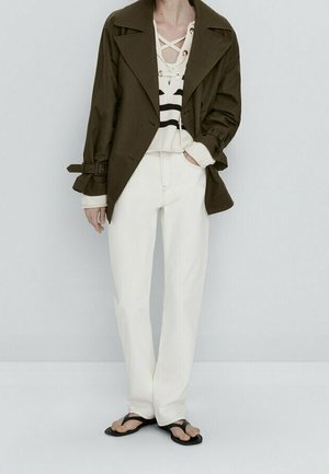 Olijfgroene trenchcoat met grote revers, gelaagd over een crèmekleurige top met zwarte strepen en een vetersluiting. Witte rechte broek en zwarte flip-flops.