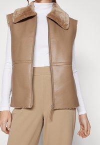 Brązowa kamizelka z sztucznej skóry z teksturowanym beżowym kołnierzem, zamkiem błyskawicznym i bez rękawów. Noszona na białym golfie.