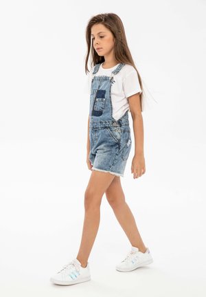 Salopette en denim bleu clair avec poignets retroussés, une poche avant, portée sur un t-shirt blanc. Baskets blanches avec des accents colorés.