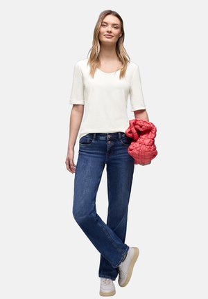 Jeune femme portant un haut blanc et un jean bleu, tenant une veste matelassée rouge pliée, debout les jambes croisées, baskets blanches.