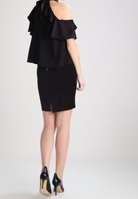 Top nero off-the-shoulder con dettagli a volant abbinato a una gonna nera aderente con spacco sul retro. La modella indossa tacchi alti neri.