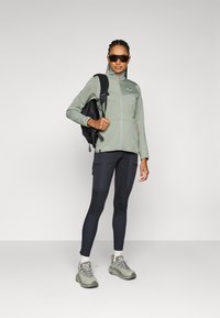 Lys grønn hettejakke med glidelås, svart ryggsekk, svarte leggings med lommer og grå sko. Solbriller fullfører det uteklare utseendet.