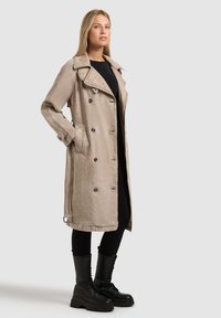 Nő bézs színű, dupla soros ballonkabátban fekete ruházaton, fekete fűzős bakancsban, zsebébe dugott kézzel áll.