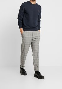 Maglione girocollo blu navy, pantaloni grigi a quadri e scarpe nere. Il maglione presenta un piccolo emblema; i pantaloni sono sartoriali con risvolti.
