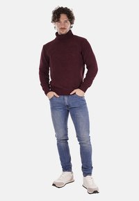 Maglione a collo alto bordeaux, con texture a coste, abbinato a jeans azzurri chiari e sneakers bianche con dettagli neri, in piedi su uno sfondo neutro.