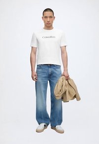 Άνδρας που φοράει λευκό t-shirt Calvin Klein, φαρδιά μπλε τζιν, λευκά αθλητικά παπούτσια, κρατώντας ένα μπεζ σακάκι στο δεξί του χέρι.