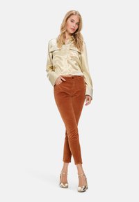 Goldfarbener Satinhemd mit spitzem Kragen und zwei Brusttaschen, kombiniert mit kastanienbraunen Cordhosen. Das Model trägt metallische Riemchen-High Heels.
