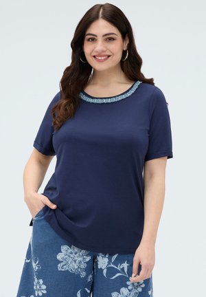 Kobieta o długich ciemnych włosach, ubrana w granatowy krótki top z ozdobnym dekoltem i szerokie, niebieskie spodnie w kwiaty, stojąca z jedną ręką w kieszeni.