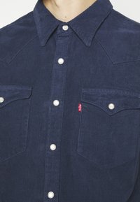 Tummansininen samettipaita, jossa on terävä kaulus, hopeiset nepparit, kaksi rintataskua ja pieni punainen Levi's-tagi.
